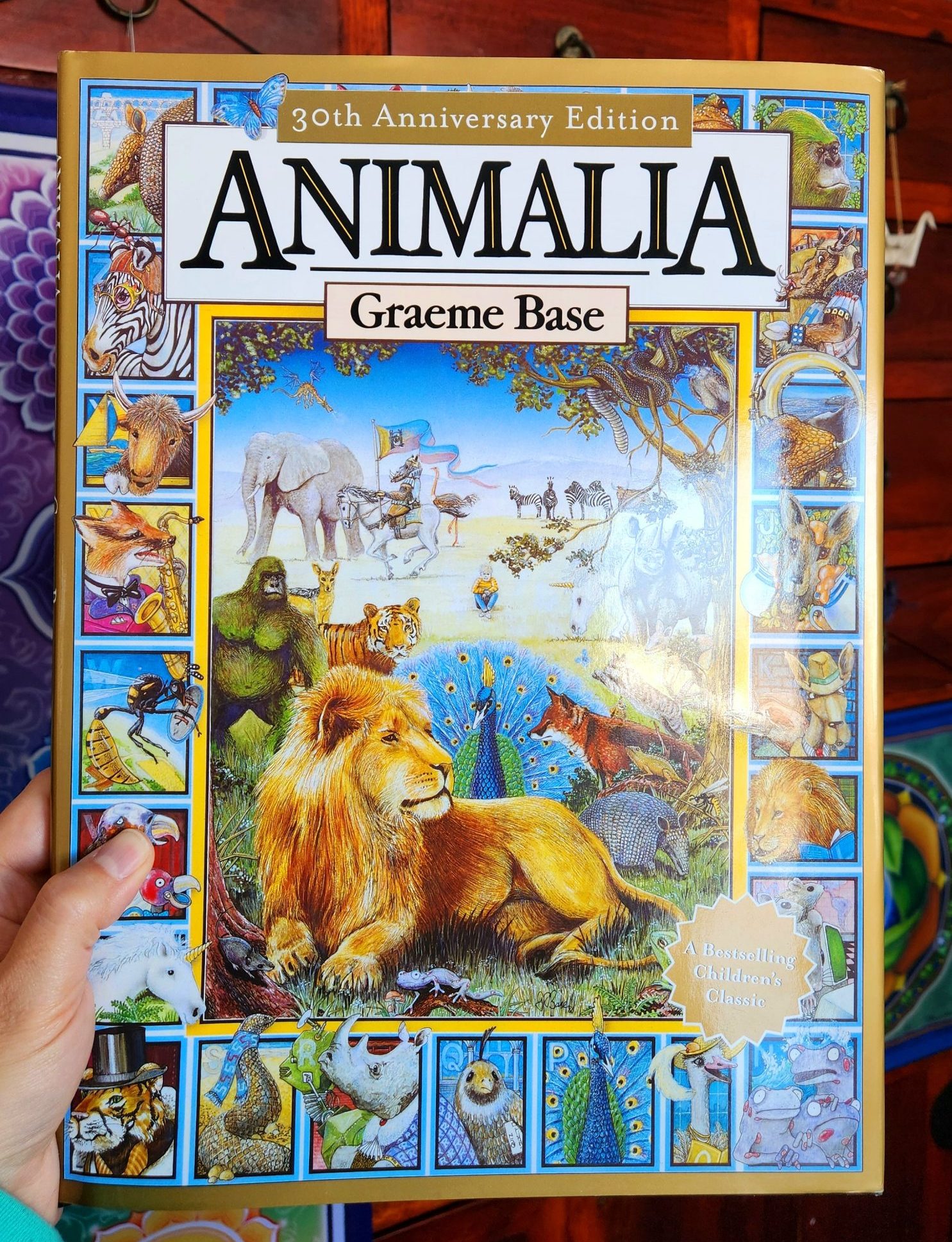 Lya's hand holding up the book Animalia by Graeme Base in front of a cherrywood piece of furniture