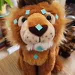 Stuffed lion sitting on a desk with small blue square stickers on the EFT points of his face